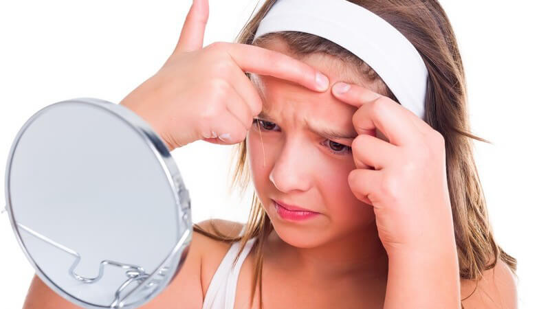 Teenager mit weißem Stirnband steht vor einem Kosmetikspiegel und drückt sich einen Pickel auf der Stirn aus