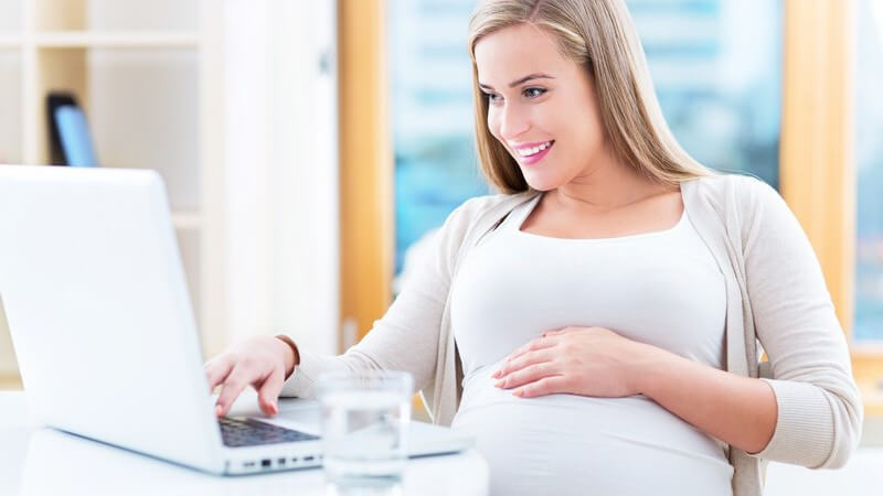 Schwangere Frau sitzt mit einem Glas Wasser lächelnd vor einem Laptop