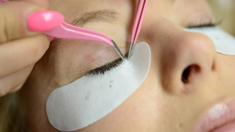 Kosmetikerin nimmt mit rosa Pinzetten eine Wimpernverlängerung bei Kundin vor, weiße Pads unter den Augen