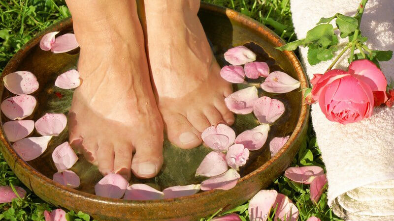 Fußbad in Schale mit Rosenblütenblättern auf Wiese, Rose und Handtuch daneben