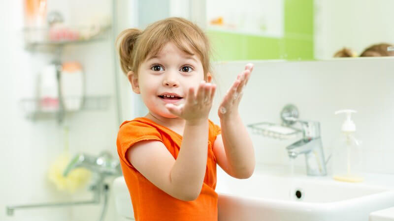 Kleines Mädchen in orangenem Shirt steht im Badezimmer am Waschbecken und wäscht sich die Hände