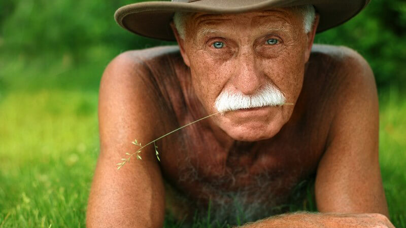 Braungebrannter alter Mann mit weißem Schnurrbart, Hut, nacktem Oberkörper liegt auf Bauch auf Wiese