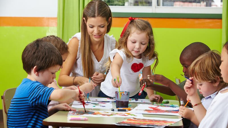 Kindergärtnerin sitzt mit Kindern am Tisch, die mit Wasserfarbe Bilder malen