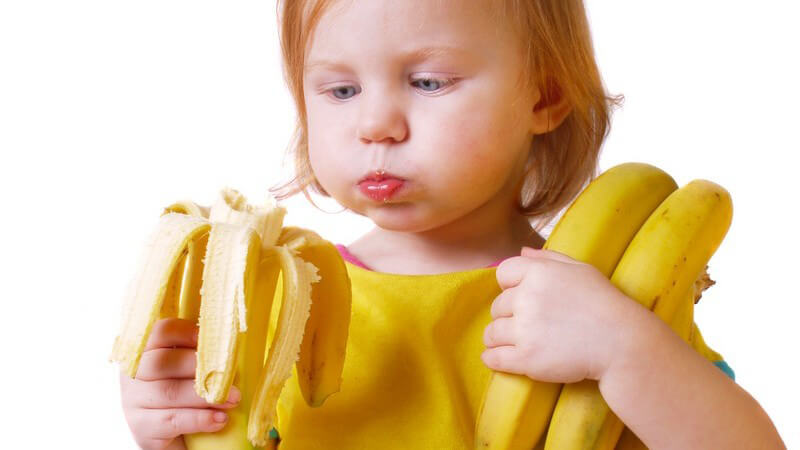 kleines rothaariges Mädchen in gelbem T-Shirt hält Bananen im Arm und isst eine Banane