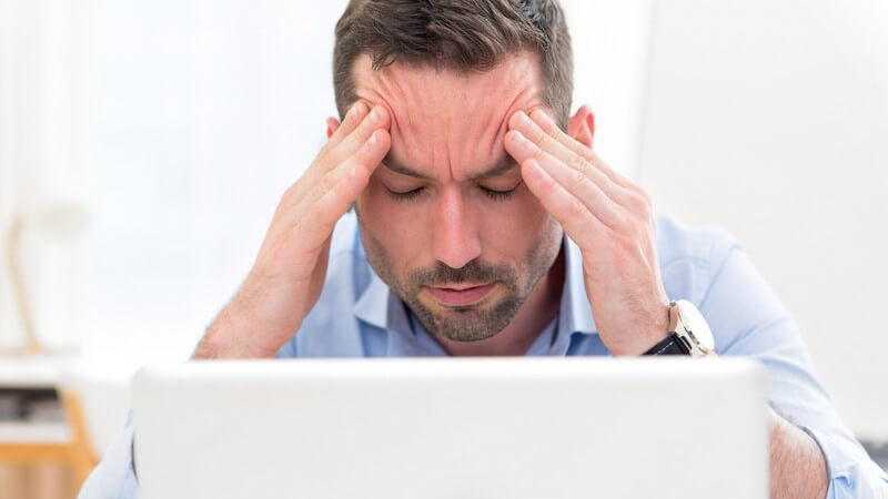 Geschäftsmann in blauem Hemd sitzt vor weißem Laptop und fasst sich vor Kopfschmerzen an die Stirn