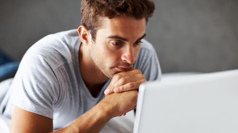 Mann liegt auf Bett und schaut auf Notebook-Bildschirm