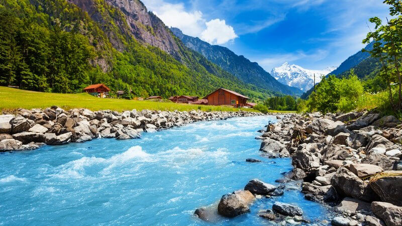 Fließender Bach/Fluss in einem schweizer Gebirge, im Hintergrund Häuser und Berge
