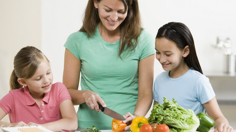 Zwei Mädchen schauen einer Frau beim Gemüseschneiden zu