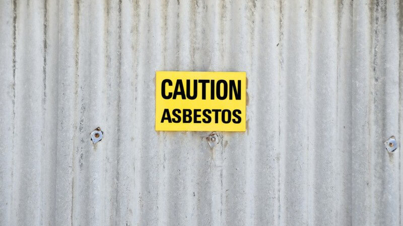 Gelbes Schild mit Aufschrift "Achtung, Asbest" auf Blechwand