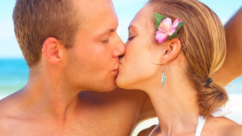 Junges Paar küsst sich vor blauem Meer, sie hat rosa Blüte im Haar