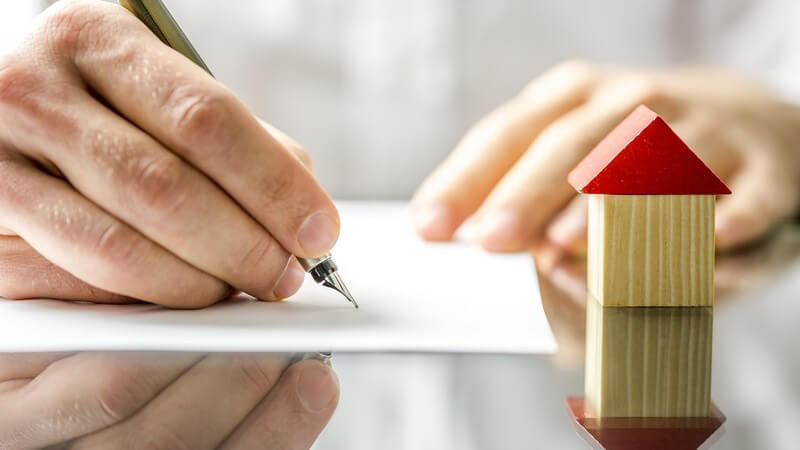 Vertragsunterschrift mit einem Füller, daneben ein kleines Holzmodell eines Hauses mit rotem Dach