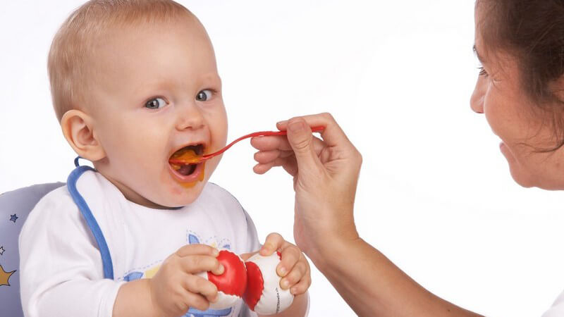 Baby wird mit Löffel gefüttert, hält zwei kleine Bälle in Händen