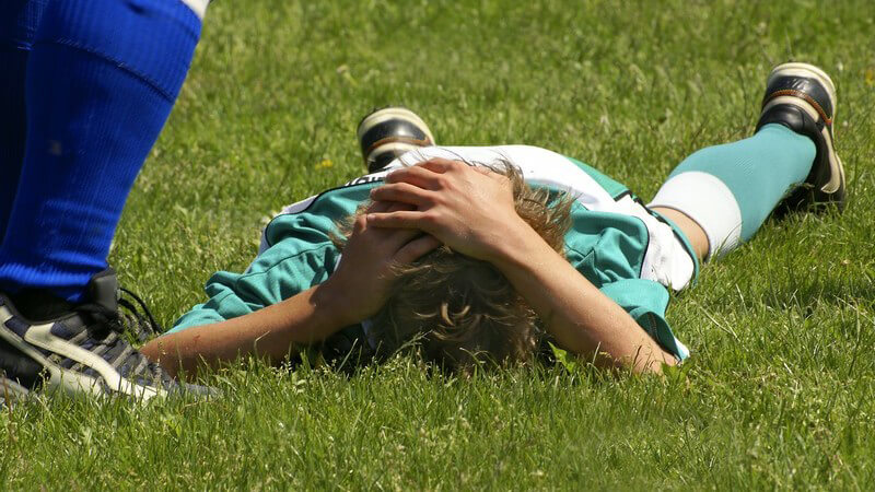 Junge in Fußballtrikot liegt auf dem Bauch auf dem Rasen, die Hände auf dem Kopf