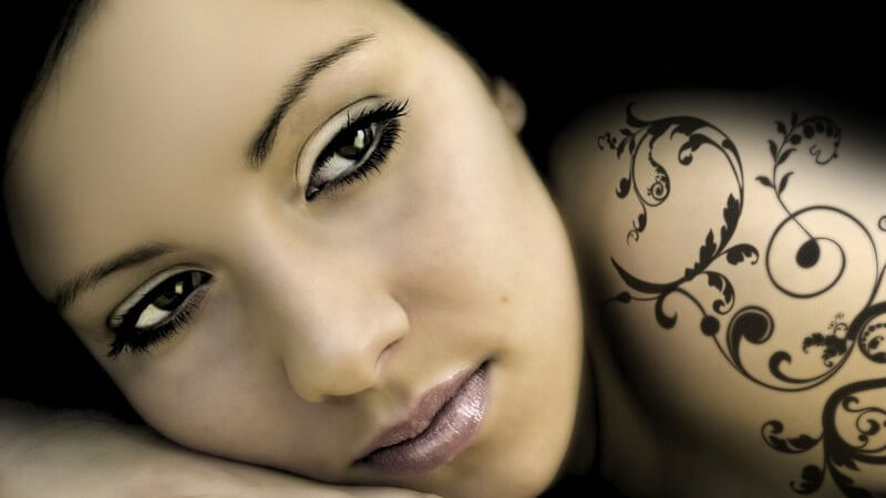 Nahaufnahme junge dunkelhaarige Frau hat Gesicht auf Hand gestützt, am Arm Grafik Tattoo, geschwungene Linien