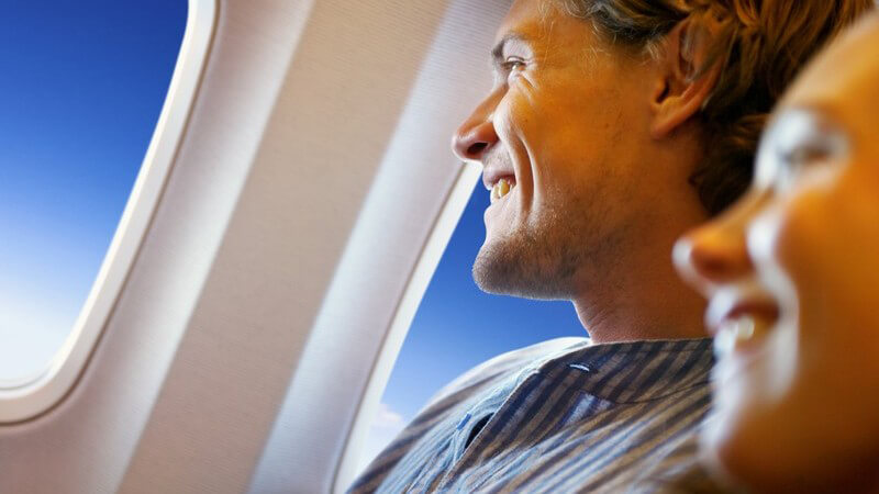 Junger lächelnder Mann auf Fensterplatz in Flugzeug, neben ihm lächelnde junge Frau