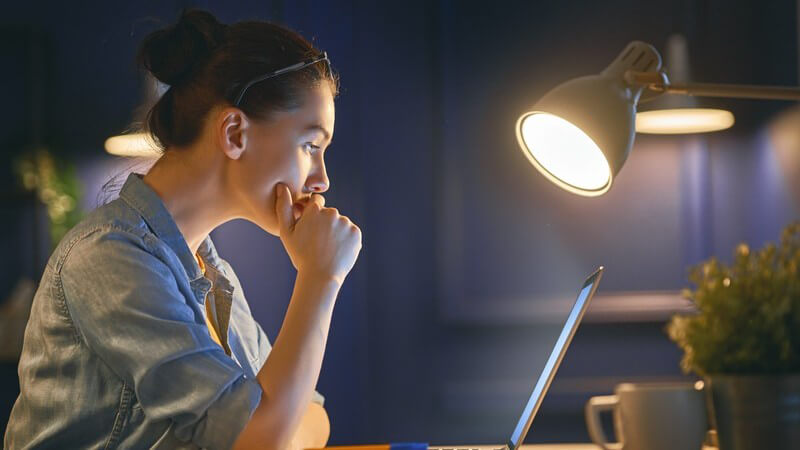 Dunkelhaarige Frau mit Brille im Haar arbeitet im Licht einer Schreibtischlampe am Laptop