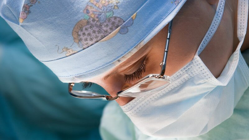 Ärztin mit Brille und hellblauer Kappe mit buntem Motiv blickt nach unten, wo sie angestrahlt wird