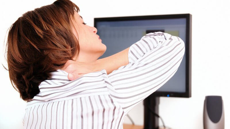 Frau sitzt vor einem Bildschirm und fasst sich vor Schmerzen an den Nacken