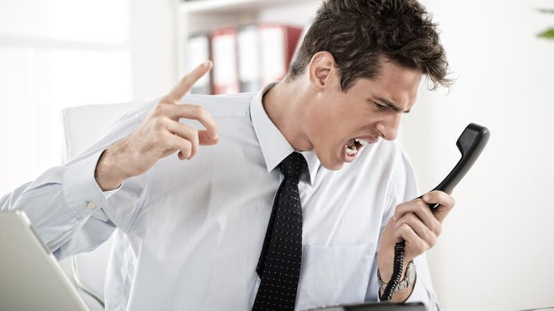 Gestresster Geschäftsmann am Schreibtisch schreit wütend in Telefonhörer rein