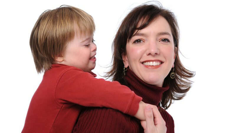 Lächelnde Mutter trägt lächelndes, behindertes Kind mit Down-Syndrom auf dem Rücken Huckepack