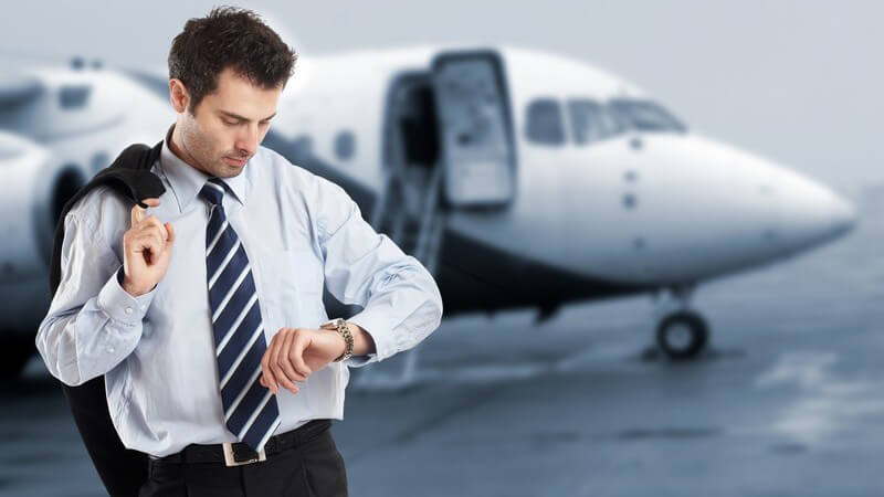 Geschäftsmann am Flughafen schaut auf Armbanduhr, hinter ihm Flugzeug