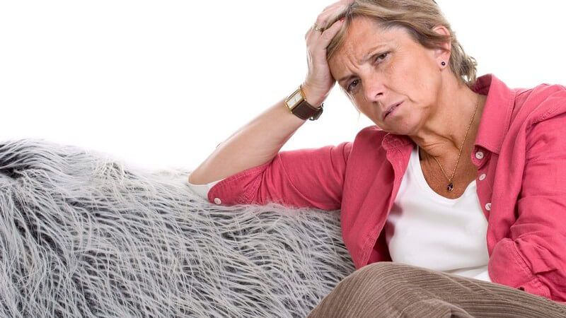 Ältere Dame in roter Bluse sitzt auf einer grauen Couch, den Kopf auf die rechte Hand gestützt