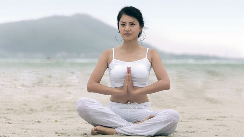 Schwarzhaarige Frau in Yogapose im Schneidersitz mit weißem Outfit am Strand