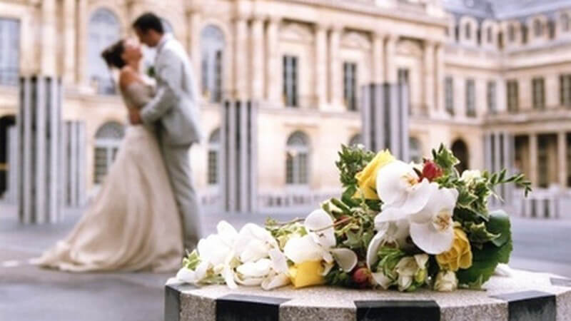 Brautpaar küsst sich vor einem Schloss, im Vordergrund ein Blumenstrauß auf einem Podest