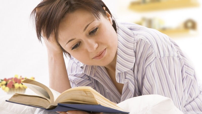 Frau liegt auf dem Bauch und liest ein Buch, die rechte Hand stützt den Kopf