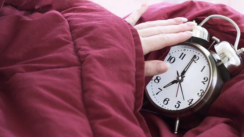 Wecker zeigt sieben Uhr, Hand schaut aus Bettdecke hervor und macht ihn aus