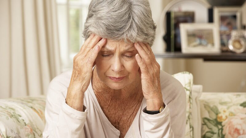 Ältere Frau mit grauen Haaren sitzt auf dem Sofa und hält die Hände an die Stirn