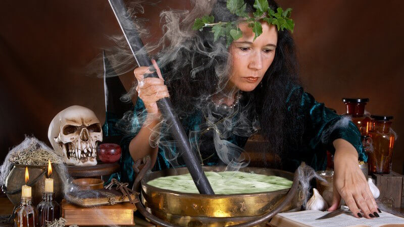 Frau als Hexe verkleidet rührt in grüner Flüssigkeit rum, umgeben von Totenkopf, Kerzen etc.