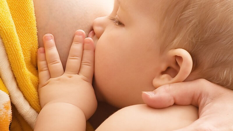 Kleines nacktes Baby wird von Brust der Mutter mit Muttermilch gestillt, hält Hand auf Brust, Mutter mit gelbem Handtuch