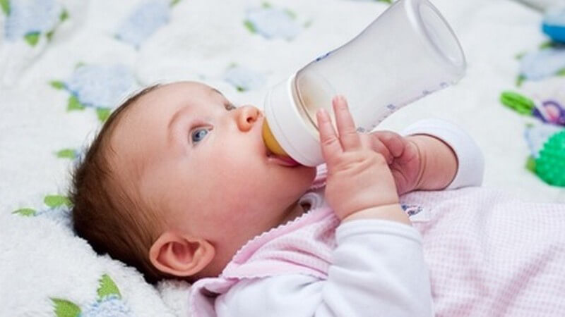 Baby im rosafarbenen Strampler mit Trinkflasche im Mund blickt nach oben
