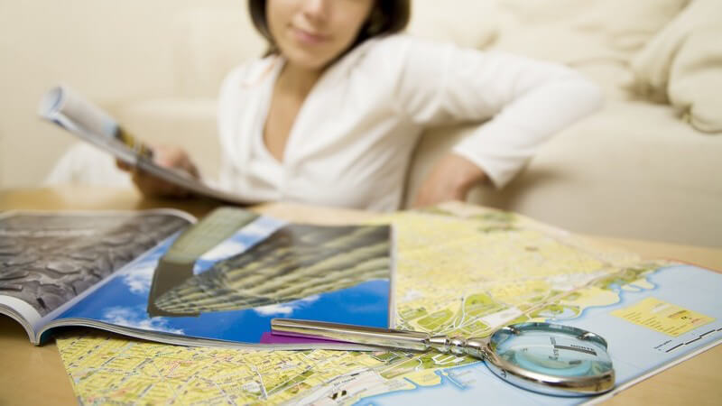 Junge Frau mit Reiseführer vorCouch, vor ihr  Landkarte für Touristen und Lupe auf Tisch