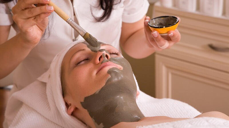 Kosmetikerin trägt einer jungen Frau eine braune Gesichtsmaske mit dem Pinsel auf