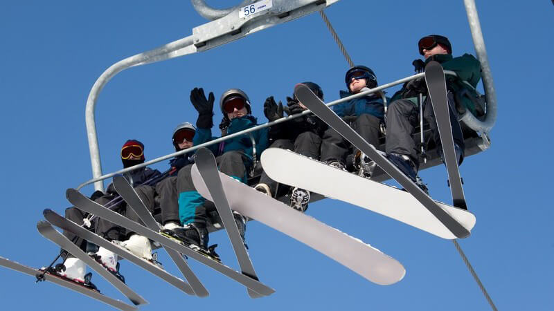 Sechs Ski-/Snowboardfahrer auf Sessellift unter blauem Himmel