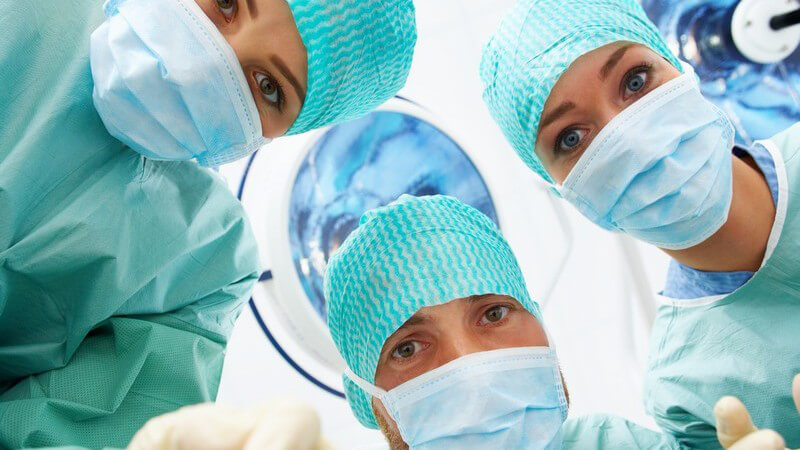 Perspektive eines auf OP-Tisch liegenden Patienten, drei Ärzte in grünen Kitteln und Mundschutz schauen auf ihn hinab