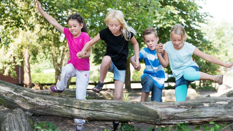 Vier Kinder springen händchenhaltend über einen Baumstamm
