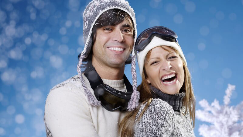 Junges lachendes Paar in Skiausrüstung, er steht hinter ihr, blauer Hintergrund mit Schneeflocken