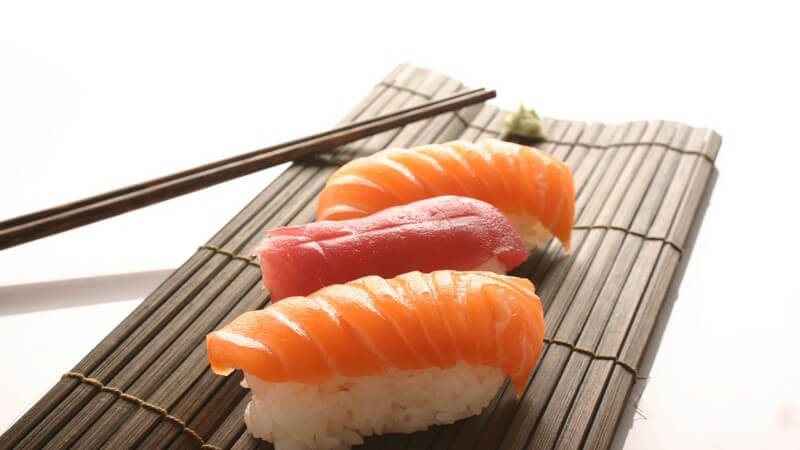 Nahaufnahme drei Happen Sushi auf Bastmatte, daneben Stäbchen
