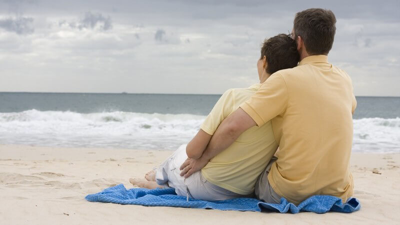 Pärchen sitzt auf einem blauen Handtuch am Strand und schaut aufs Wasser