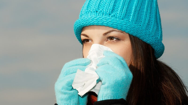 Junge, dunkelhaarige Frau mit türkiser Mütze und Handschuhen putzt sich die Nase mit Taschentuch