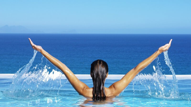 Dunkelhaarige Frau von hinten steht bis zur Brust in Pool am Meer, hebt die Arme und spritzt mit Wasser