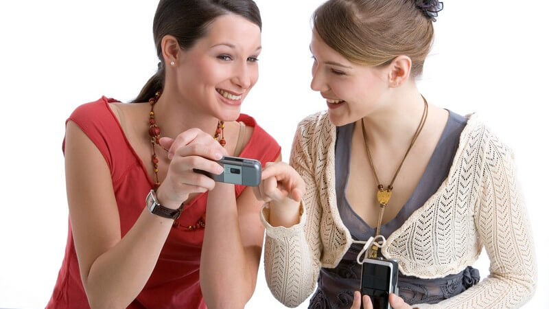 Junge Frau zeigt anderer Frau ihr Handy, Handyfoto, Unterhaltung, vor weißem Hintergrund