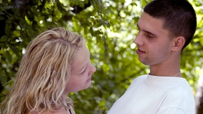 Blonde Frau und Mann mit sehr kurzen Haaren und weißem T-Shirt schauen sich im Wald intensiv an