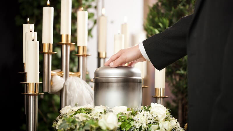 Beerdigung: Hand eines Mannes im schwarzen Anzug auf Urne, im Hintergrund weiße Kerzen