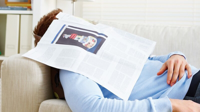 Junger Mann im Wohnzimmer auf Couch eingeschlafen, aufgeschlagene Zeitung über Gesicht