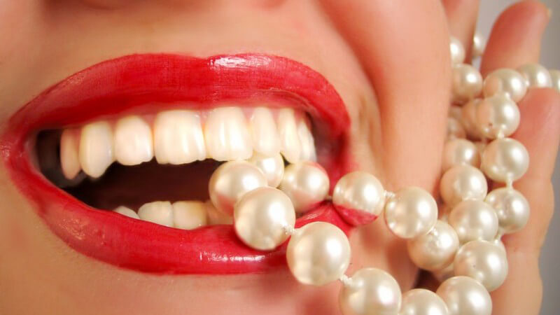 Rot geschminke Lippen einer lachenden Frau, sie beißt auf weiße Perlenkette