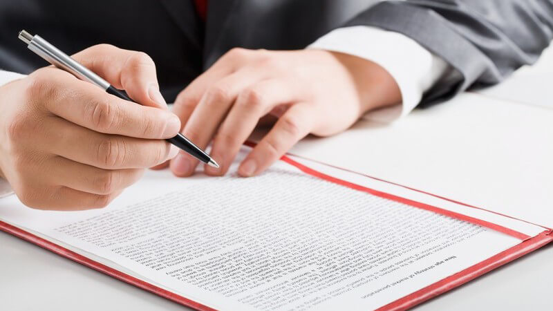 Mann im Anzug sitzt mit einem Kugelschreiber über einem Dokument im roten Schnellhefter